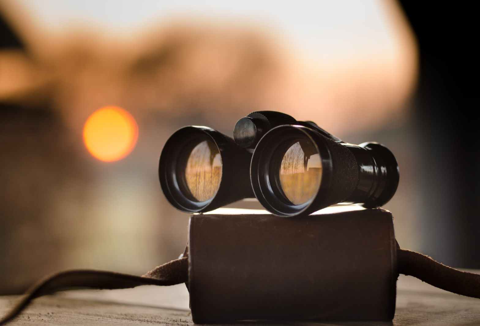 An image of black binoculars on a brown leather looking case the background it blurred but you can see the orange sun.