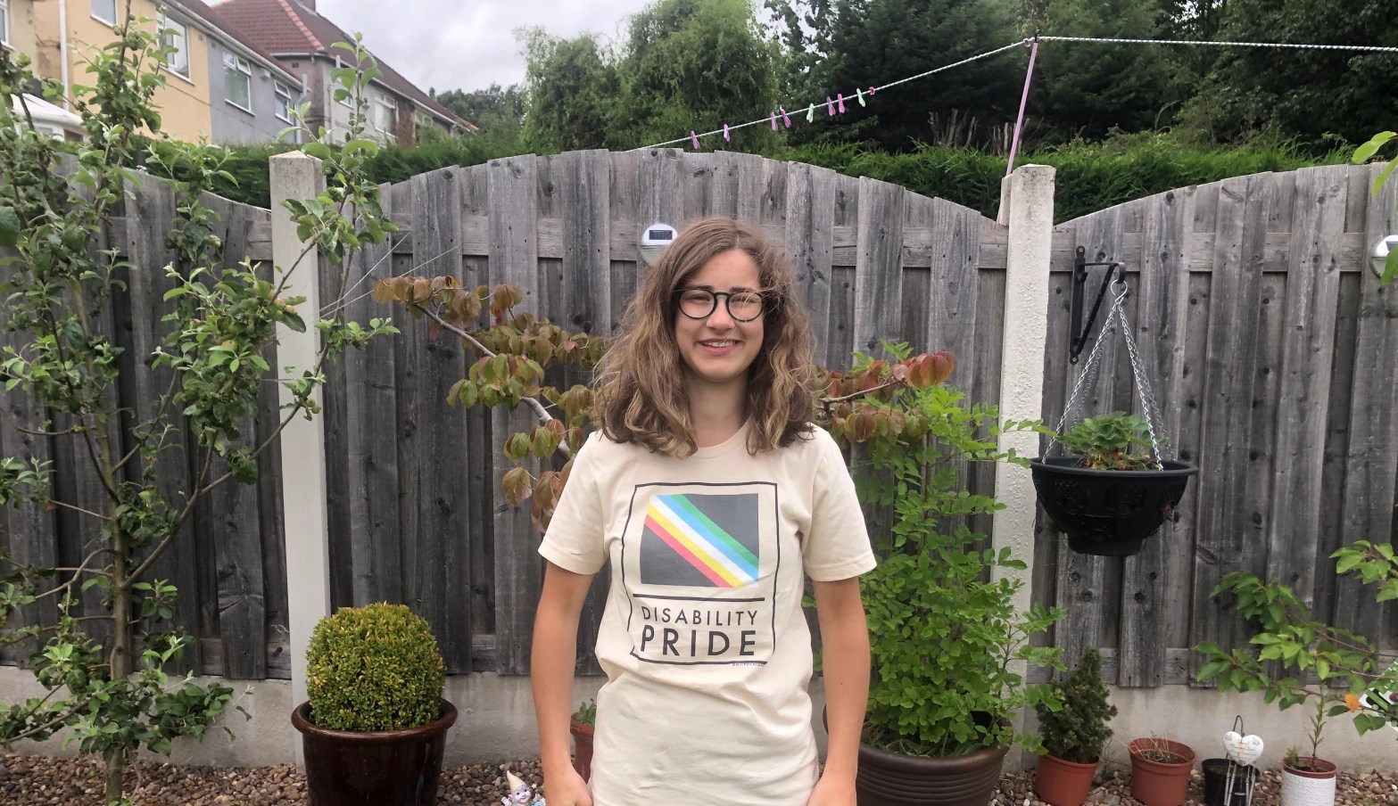 Georgia a white woman with shoulder length brunette hair stood outside in a disability pride month top.